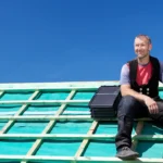 Ein zufrieden lächelnder Handwerker in Arbeitskleidung sitzt auf der Holzkonstruktion eines unfertigen Daches unter strahlend blauem Himmel.