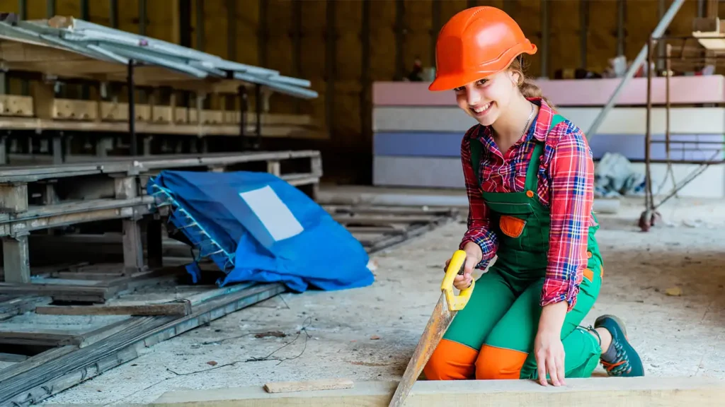 Eine lachende Person mit orangefarbenem Schutzhelm und grüner Latzhose sägt auf einer Baustelle ein Stück Holz.