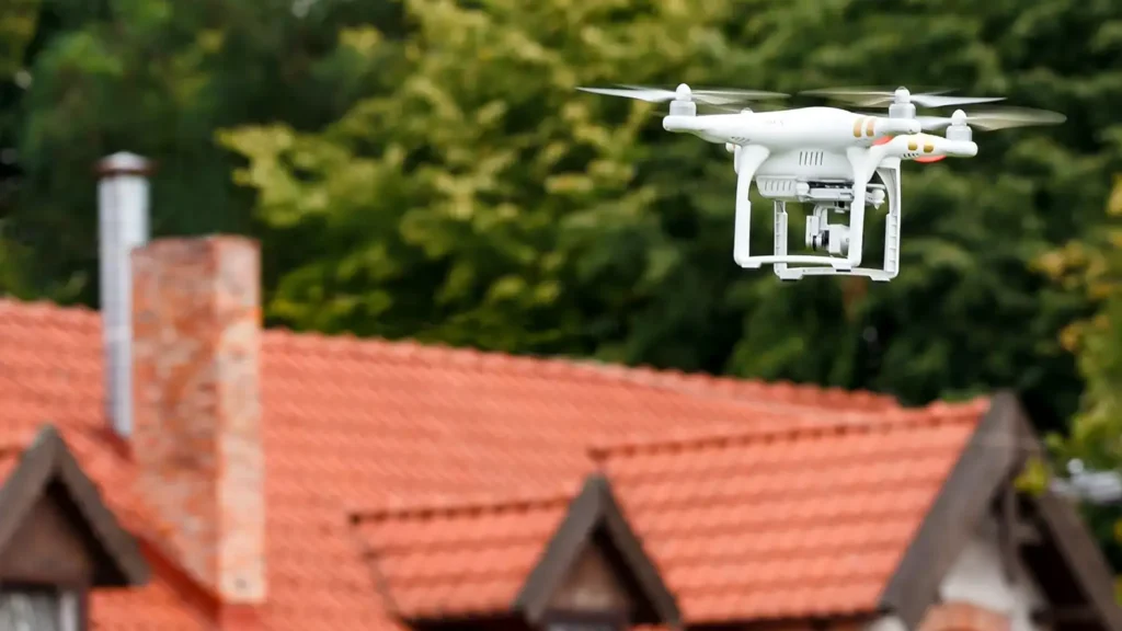 Eine weiße Quadrokopter-Drohne fliegt in der Luft; im unscharfen Hintergrund ist ein Wohnhaus mit roten Dachziegeln und grünen Bäumen erkennbar.