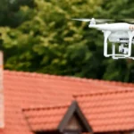 Eine weiße Quadrokopter-Drohne fliegt in der Luft; im unscharfen Hintergrund ist ein Wohnhaus mit roten Dachziegeln und grünen Bäumen erkennbar.