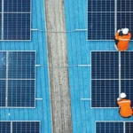 Vogelperspektive: Drei Handwerker in oranger Warnkleidung montieren Photovoltaik-Module auf einem blauen Industriedach.